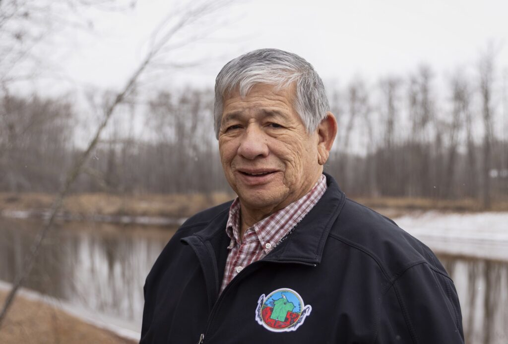 He is smiling at the camera, water and trees behind him