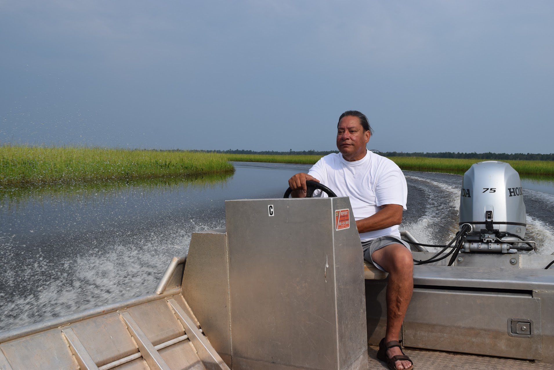 He is seated behind the wheel of a metal boat, the river bending behind him.