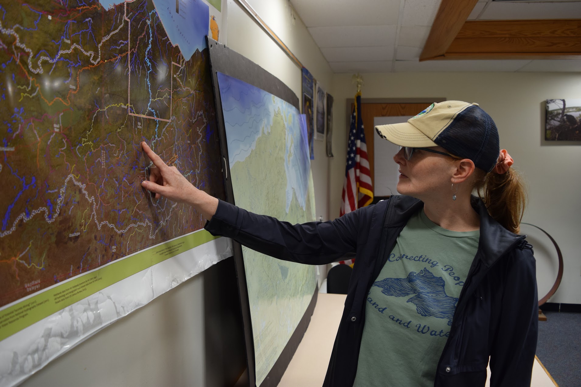 She is pointing to a map on the wall. She is wearing a shirt that says, "Connecting people, land and water"