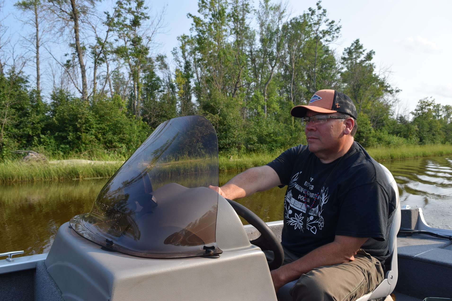 The photo shows a side view of him at the wheel, trees at the shoreline behind him