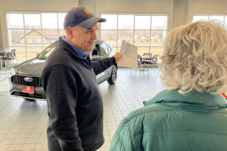 Terry Berg, who has been selling cars at Cornerstone Ford since 1987, leads a customer through the showroom at the Elk River dealership. He drives an EV and likes selling them, but finds a lot of his clients are reluctant to make the leap. Credit: Dan Gearino/Inside Climate News