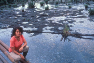 Judith Kimerling kneeling on pipelines above a drilling waste pit in the Ecuadorian Amazon in July 1990. Credit: Courtesy of Judith Kimerling